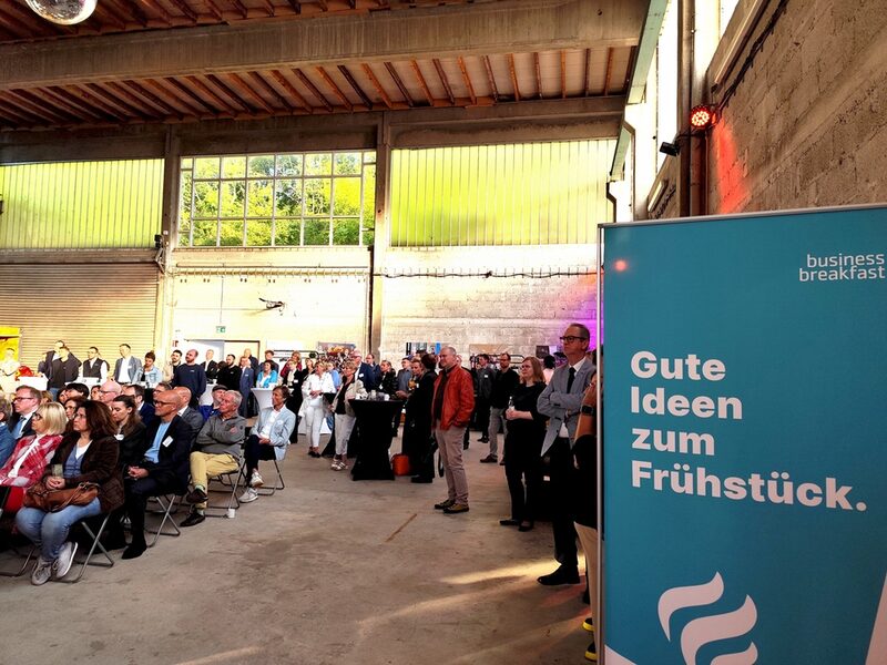 Vorne: Business Breakfast Plakat. Hinten: Gäste in der Alten Glaserei.
